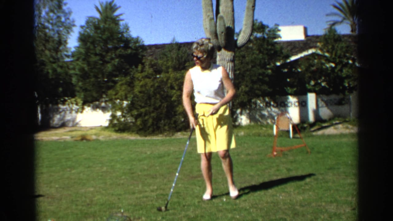 A woman golfer in 1968 Scottsdale stands over a golf ball on a residential lawn with a tall saguaro cactus and house behind her.