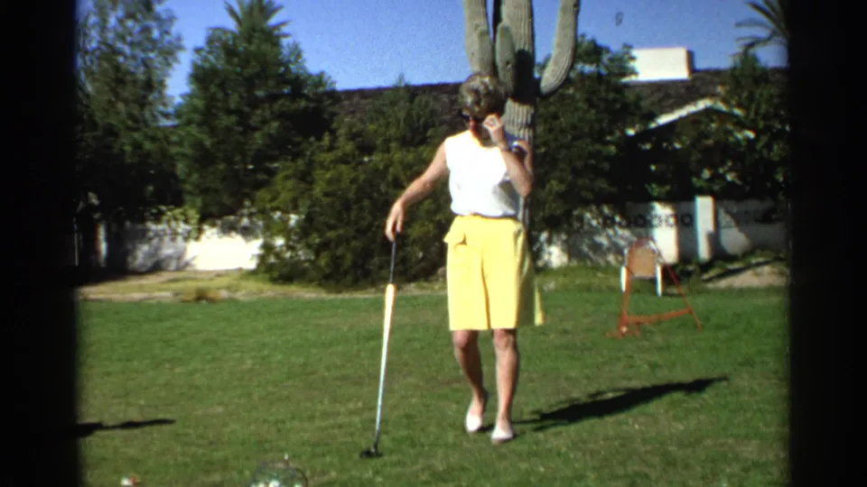The woman golfer moves near a golf ball on a Scottsdale lawn, with 1960s clothing, cactus, shrubs, and a low residential building visible.