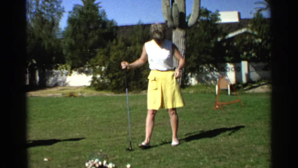 In a 1968 Scottsdale home-movie frame, the woman golfer stands near several golf balls on grass with a saguaro cactus and house behind her.