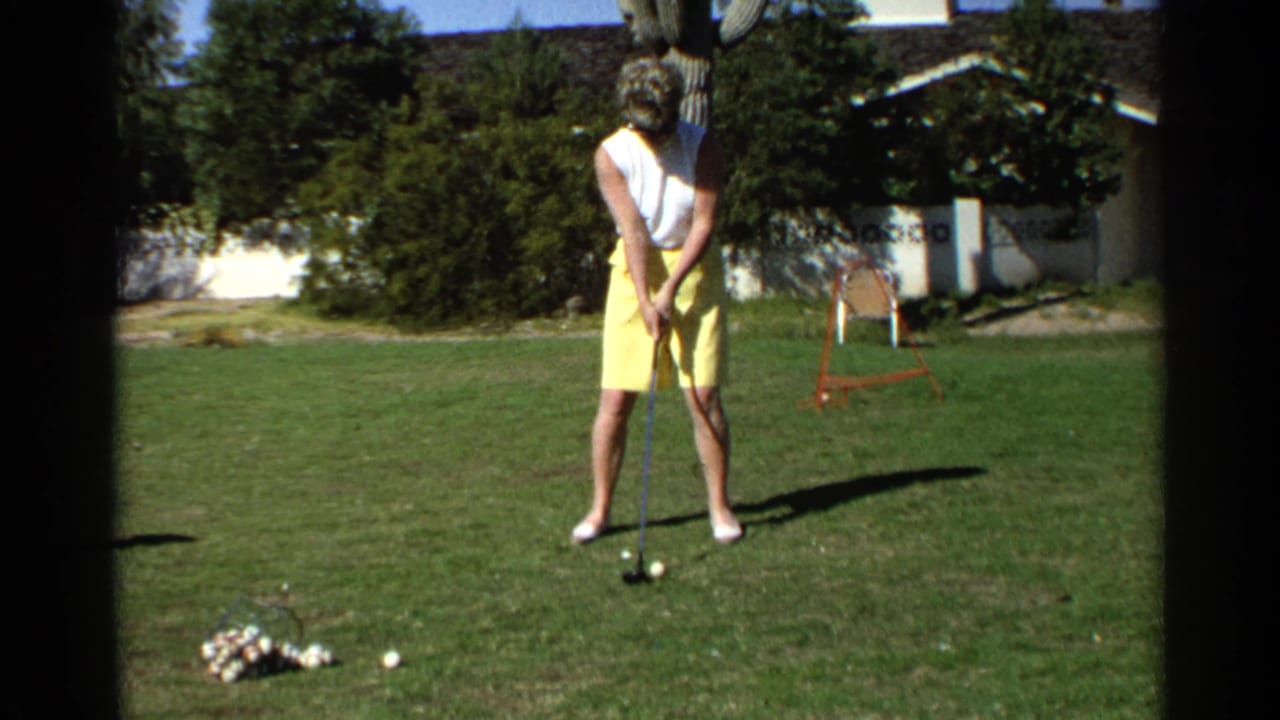 Woman stands on a backyard lawn addressing a golf ball with a club.