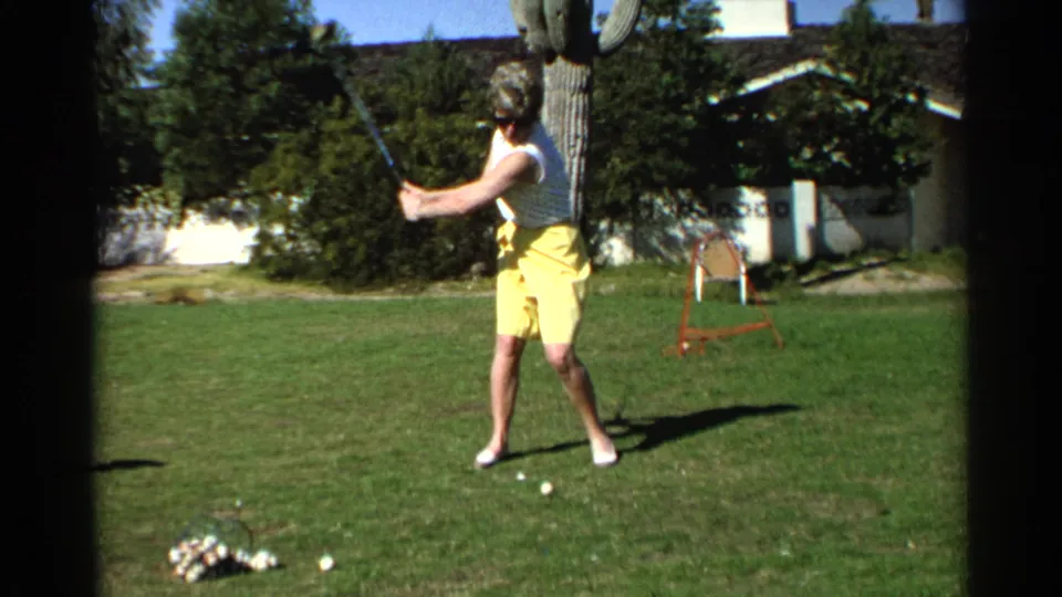 The woman starts her swing in the Scottsdale backyard, with golf balls, shrubs, and a residential exterior visible behind her.