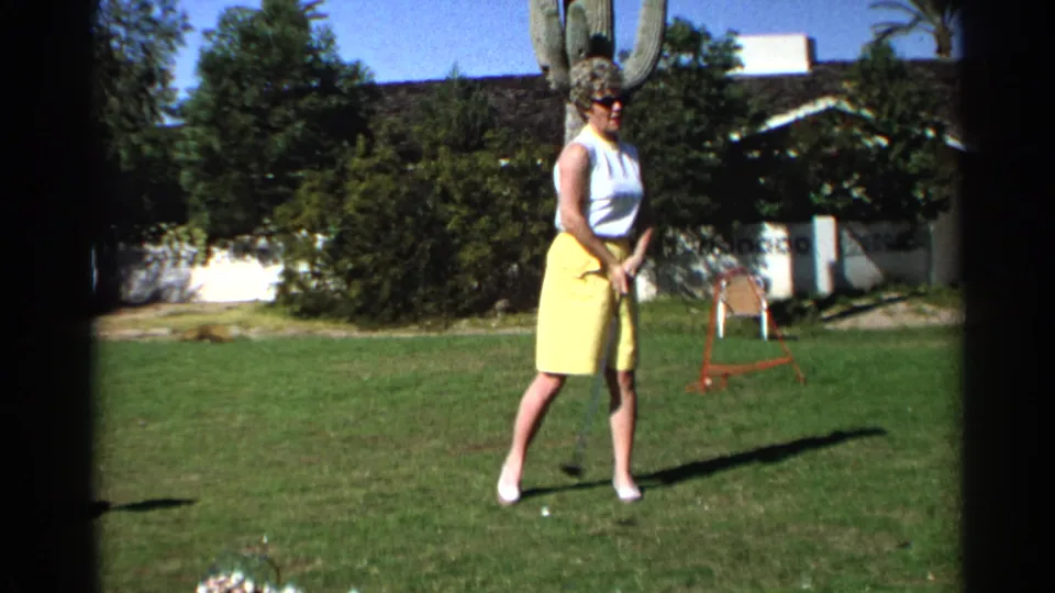 A woman in a white sleeveless top and yellow skirt stands with a golf club on a Scottsdale lawn, framed by shrubs, a cactus, and a house.