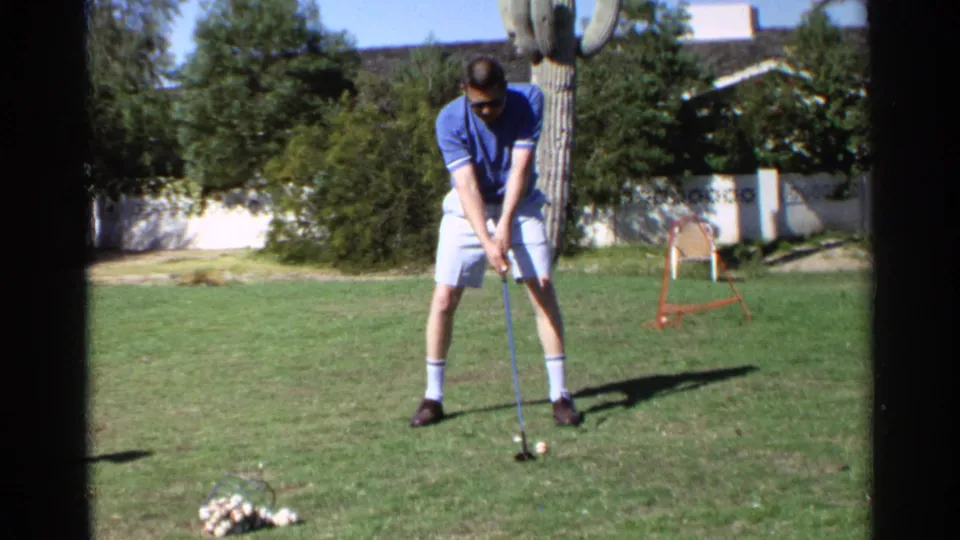 A man in a blue polo and white shorts prepares to hit a golf ball in the Scottsdale backyard, with cactus, shrubs, and a house behind him.