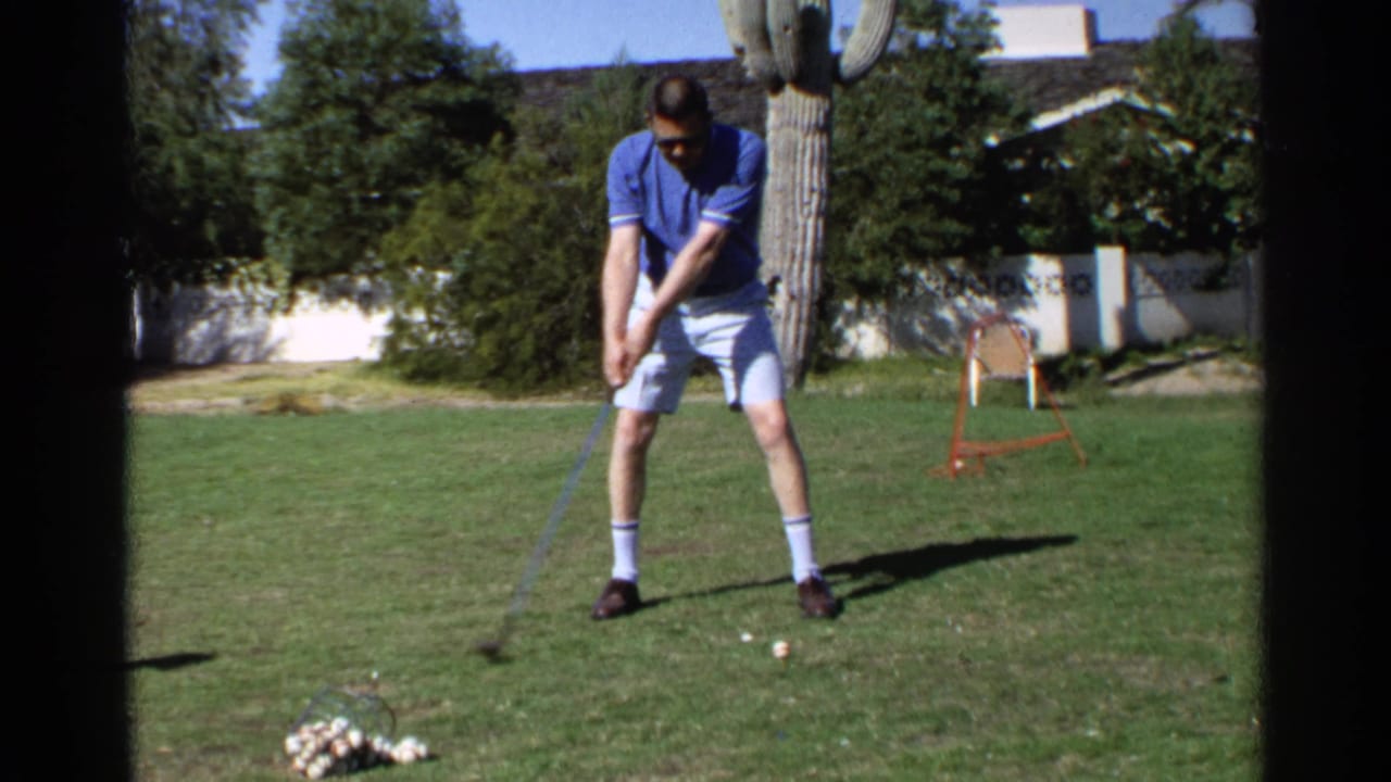 Man addresses a golf ball on a backyard lawn with a cactus behind him.