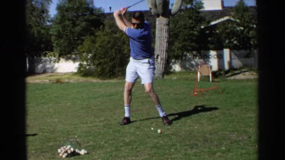 The man raises his club for a backswing in a Scottsdale backyard, with cactus, shrubs, and a patio chair in view.