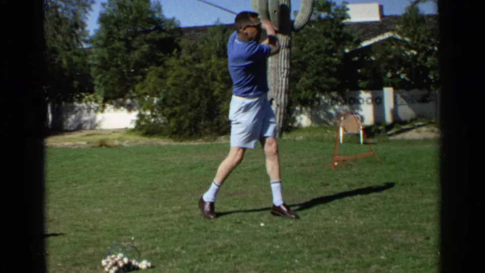 A man completes a golf swing in the 1968 Scottsdale home-movie scene, standing on grass near a cactus and patio chair.