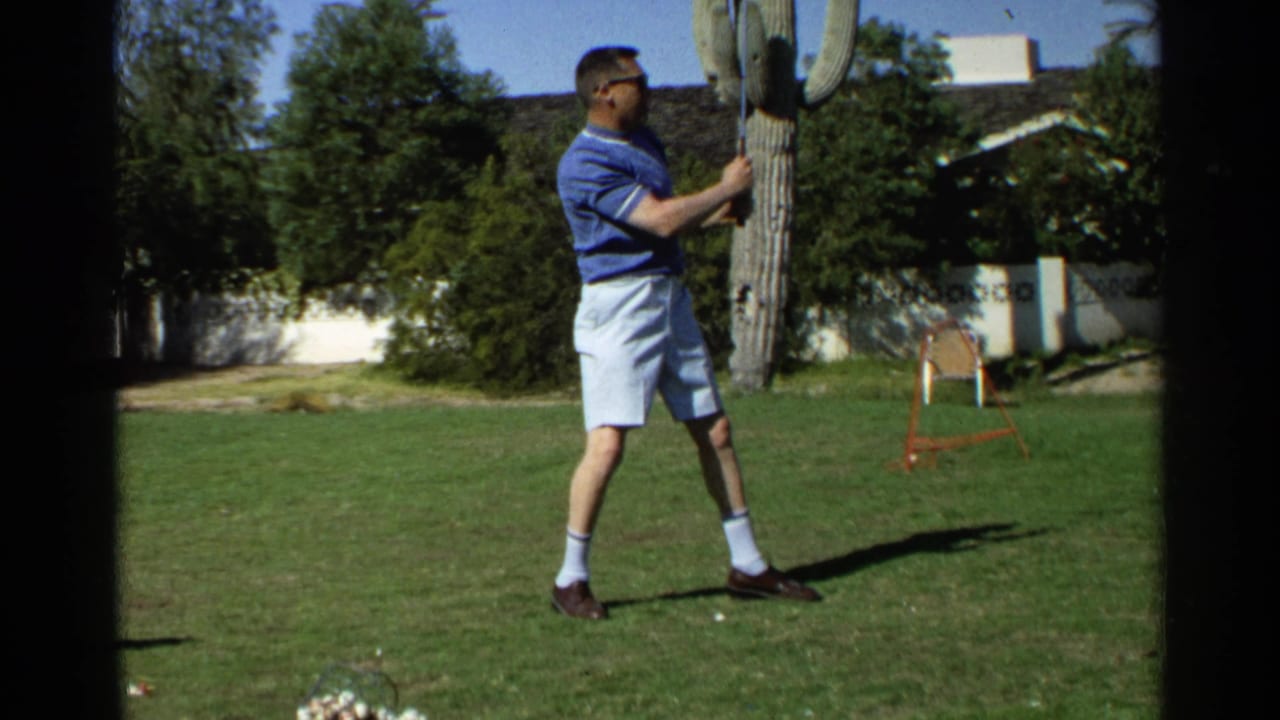 Man in a blue polo shirt swings a golf club on a sunny lawn near a saguaro cactus.