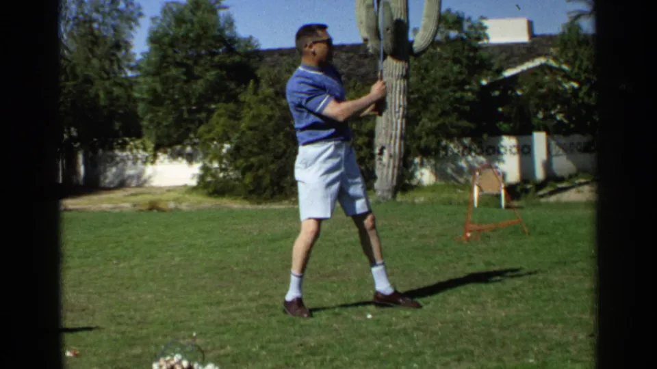 In Scottsdale, Arizona, 1968 footage, a man in a blue polo and white shorts swings a golf club on a grassy lawn beside a saguaro cactus.