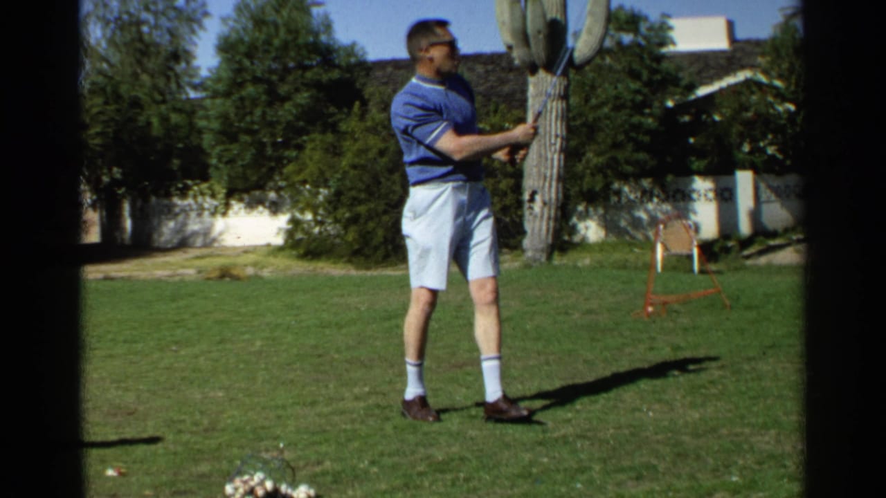 A man in 1968 Scottsdale home-movie footage holds a golf club on a lawn, with desert landscaping and a white wall in the background.
