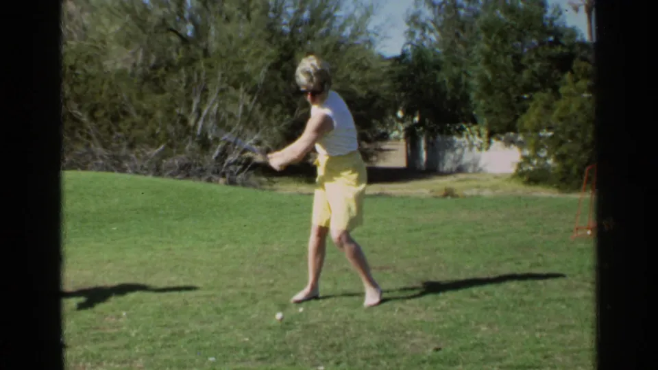 A woman in 1968 Scottsdale home-movie footage swings a golf club on a sunlit lawn bordered by trees and a low wall.