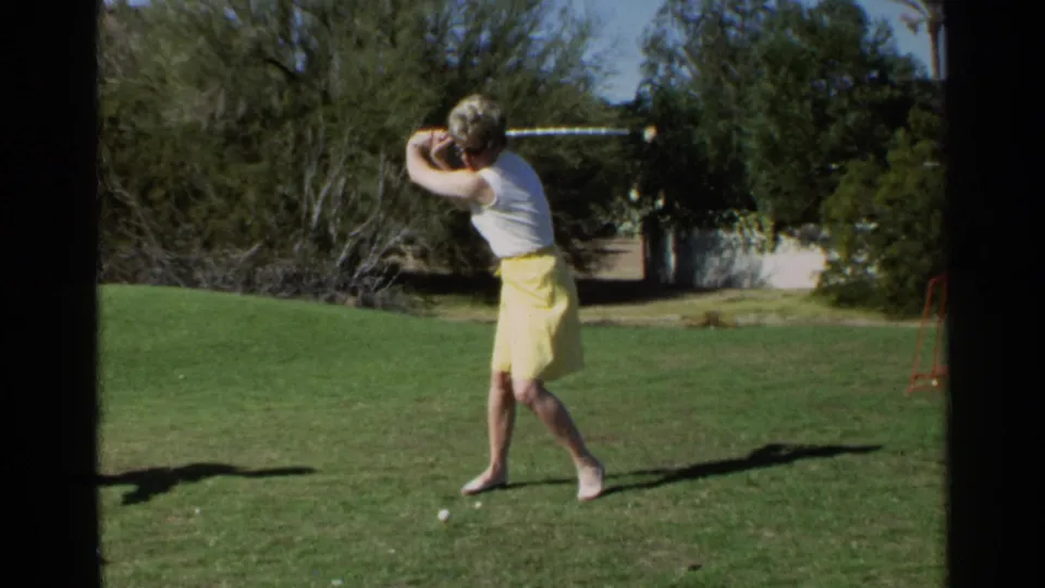 In Scottsdale, Arizona, 1968, a woman in a yellow skirt and white top prepares to strike a golf ball on a sunlit lawn.