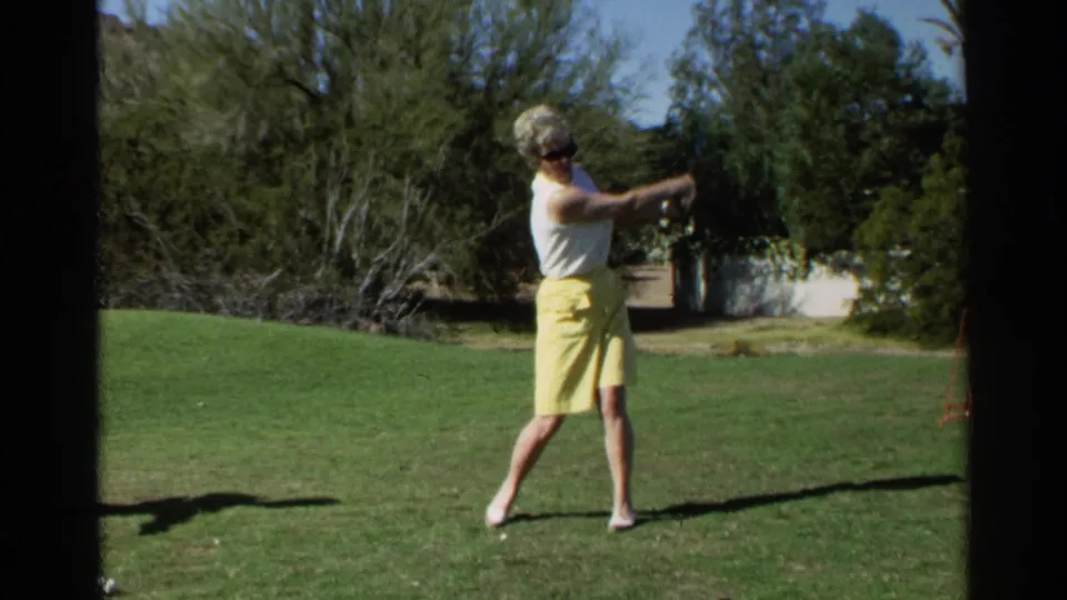 The woman golfer completes the strike in a 1968 Scottsdale home-movie frame, with lawn, trees, and a white wall in the background.