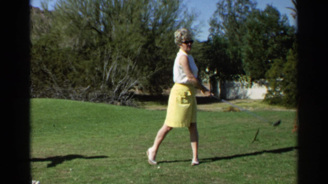 Woman in sunglasses stands on a grassy golf area holding a club.