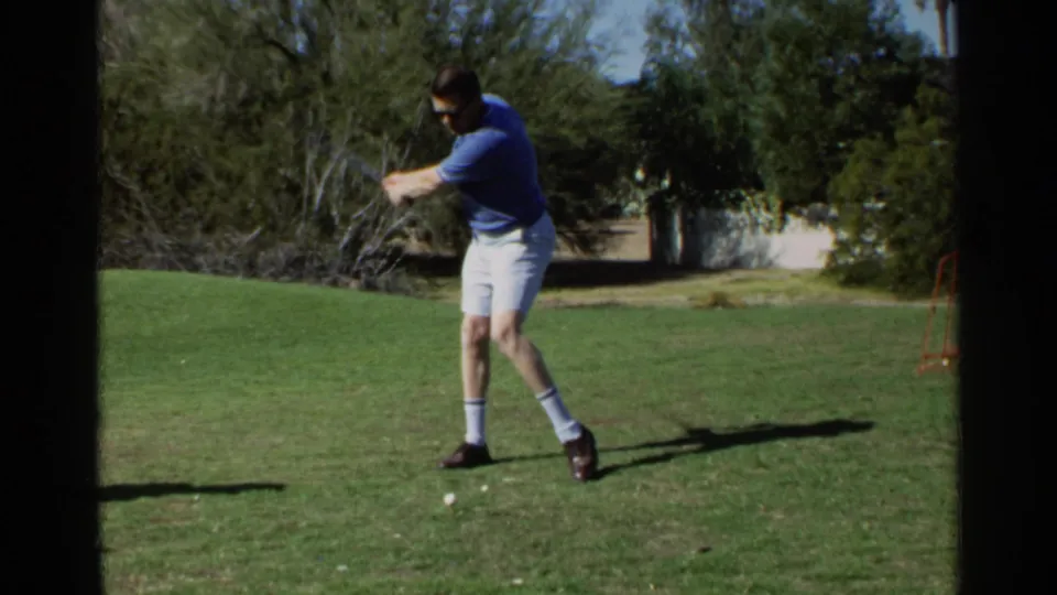 Adult man in a blue polo shirt and white shorts follows through with a golf swing in 1968 Scottsdale home-movie footage.