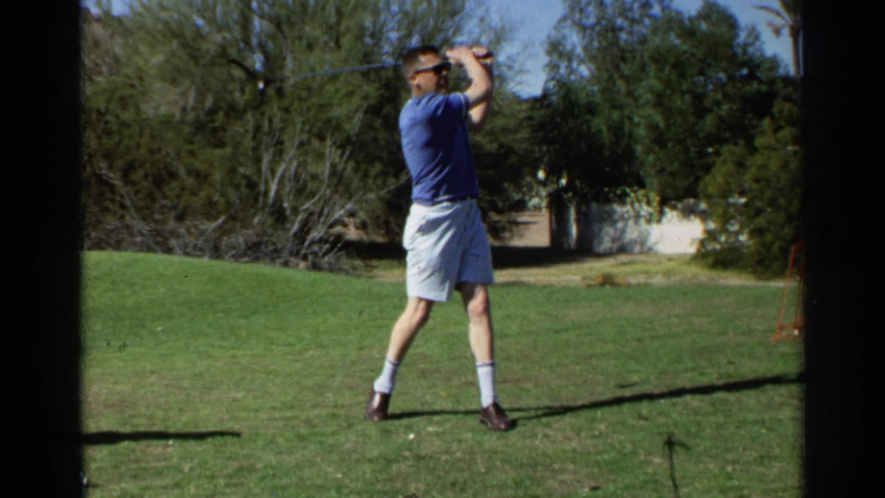 Man in sunglasses raises a golf club on a sunny lawn with desert trees behind him.