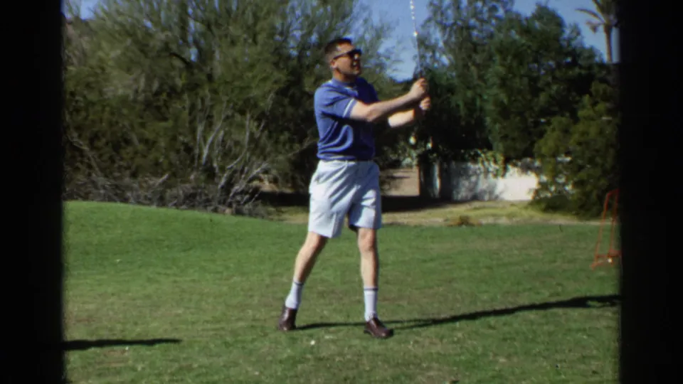 A man in a blue polo and sunglasses holds his golf club after swinging on a sunny Scottsdale lawn in 1968 home-movie footage.
