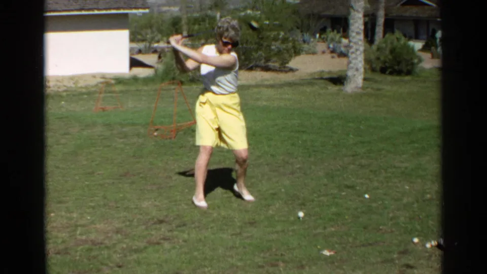 In Scottsdale, Arizona, 1968, a woman in a white sleeveless blouse and yellow skirt sets up a golf swing on a sunny lawn.