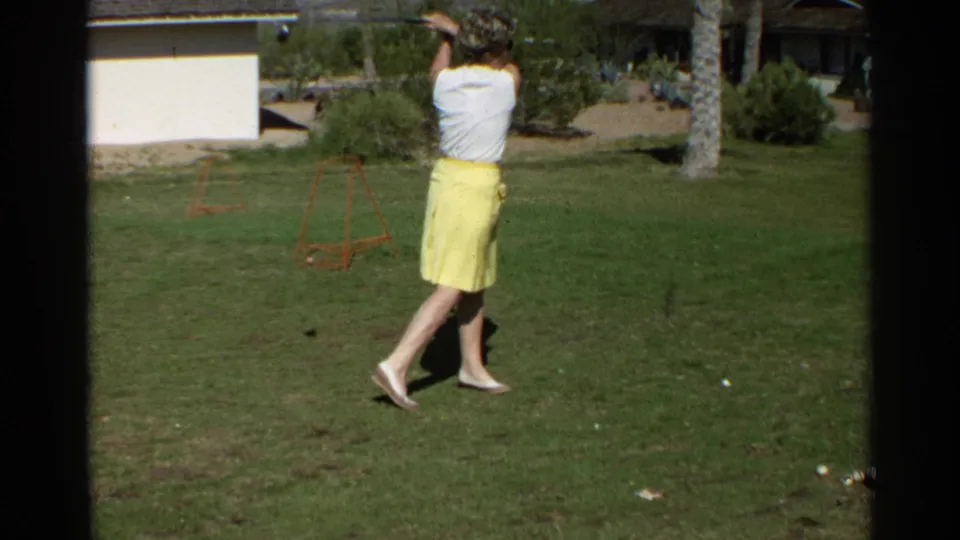 The woman follows through on a golf swing in the 1968 Scottsdale home-movie footage, with lawn, palms, and a house behind her.