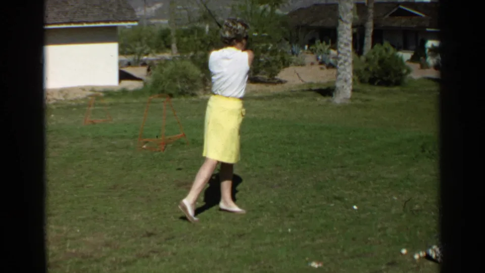 Seen from behind, the woman completes her golf swing on the Scottsdale lawn, with shrubs, palms, and a house in the background.