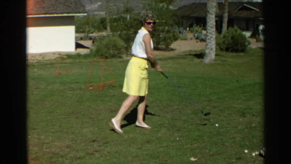 After the swing, the woman lowers the golf club and turns slightly on the Scottsdale lawn, with golf balls and residential landscaping visible.