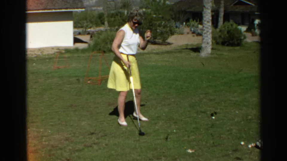 The woman pauses with one hand raised and a club grounded near a golf ball in the 1968 Scottsdale home-movie scene.