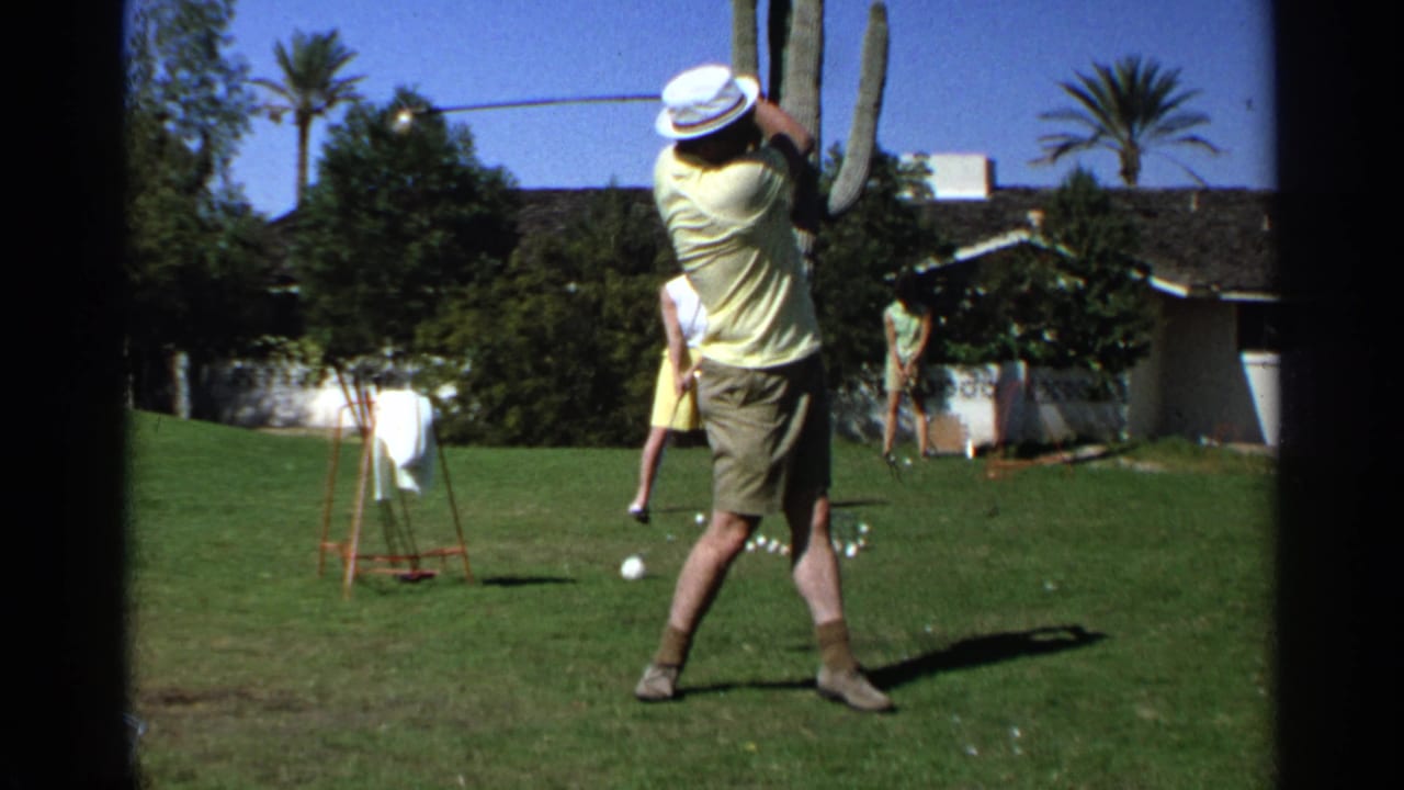 Man in a white hat swings a golf club on a grassy backyard range.