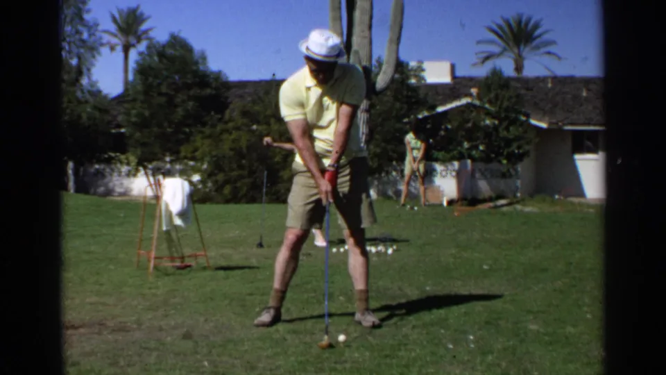 The man in a yellow shirt and green shorts prepares to strike a golf ball on a Scottsdale backyard lawn, with palms, a cactus, and another adult behind him.