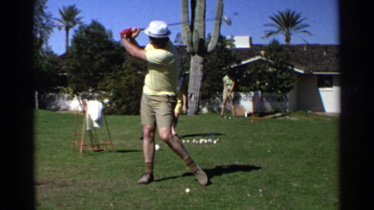 Man in a white hat begins a golf swing on a sunny lawn with a cactus and house behind him.