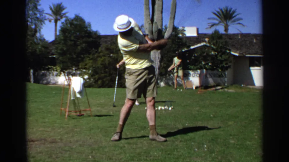 The golfer follows through on a lawn in Scottsdale, Arizona, with a saguaro cactus, palm trees, house, and other people visible behind him.