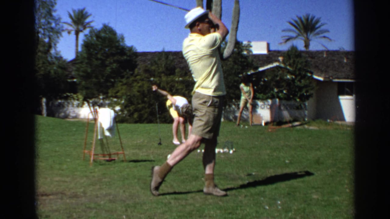 Man in a yellow shirt follows through on a golf swing on a sunny lawn.