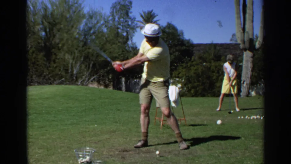 A man swings a golf club on a Scottsdale lawn as a woman in a yellow dress stands near golf balls and desert planting.