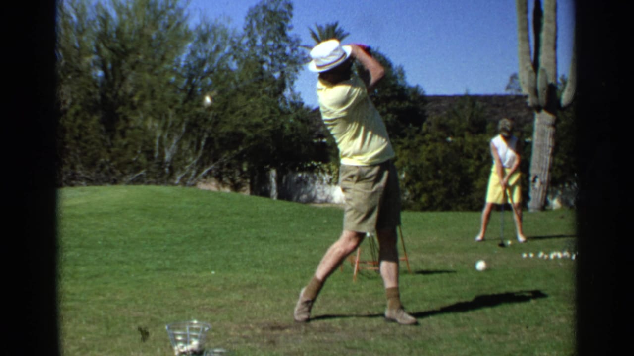 Man in a yellow shirt finishes a golf swing on a sunny grass range.