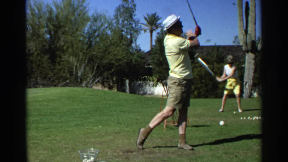 The man pauses in his follow-through on a bright Scottsdale golf range, with another adult golfer and desert vegetation behind him.