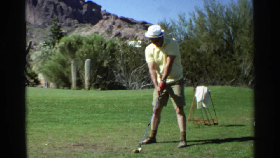 A man in a yellow shirt and white hat stands over a golf ball on a Scottsdale course, seen in 8mm home-movie footage.