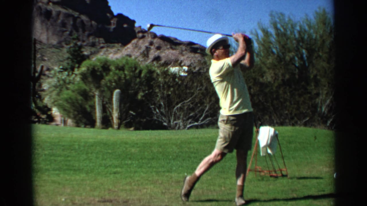 Adult golfer in a white hat follows through on a green fairway with desert plants behind him.