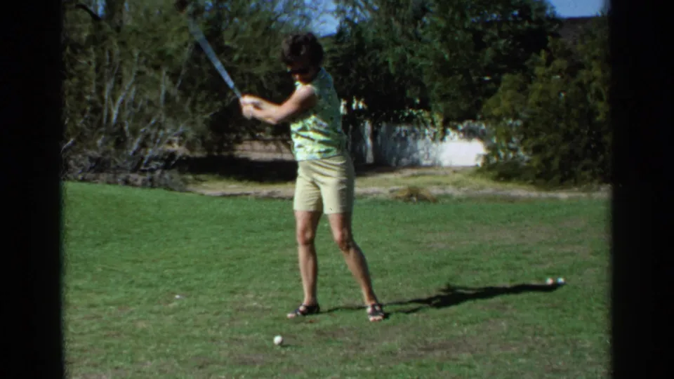 1968 Scottsdale frame of a woman finishing her golf swing, with grass, shrubs, and course trees behind her.