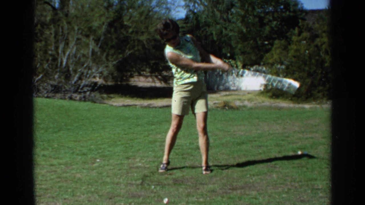 Woman in sunglasses swings a golf club on grass with trees behind her.