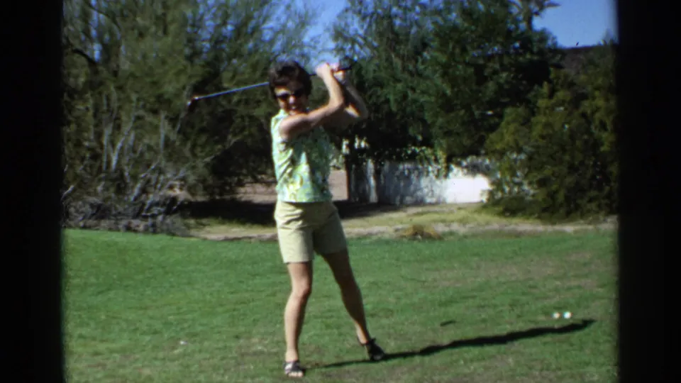 A woman in sunglasses stands with her golf club raised during a 1968 Scottsdale home-movie golf practice moment.