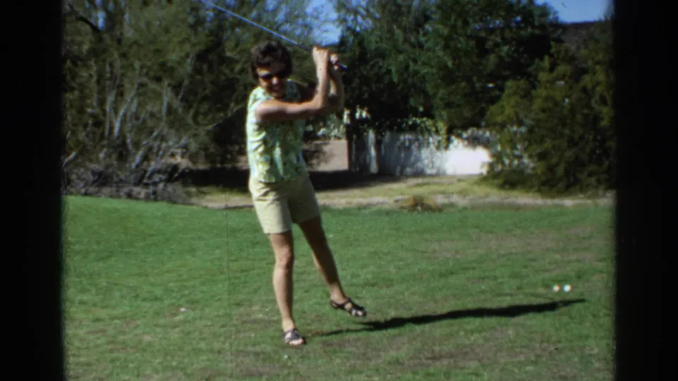 Adult woman in a green patterned top follows through on a golf swing in a sunlit Scottsdale yard in 1968.