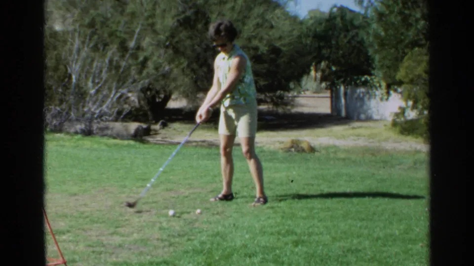 Adult woman prepares to hit a golf ball on a grassy yard in 1968 Scottsdale home-movie footage.