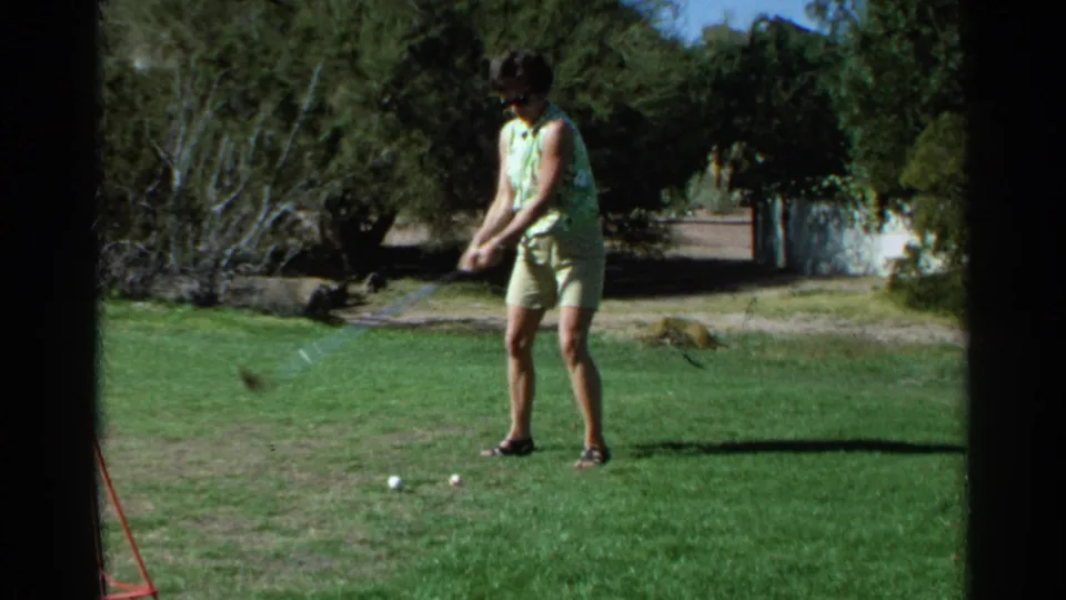The woman prepares near two golf balls on a sunny Scottsdale course, framed by trees and a pale wall in 1968 8mm footage.