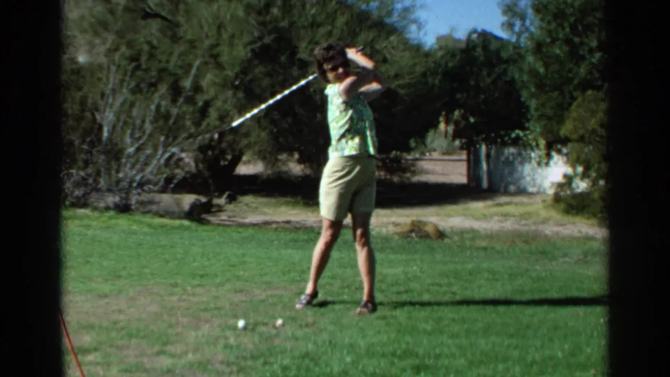 The woman finishes a golf swing with the club high over her shoulder on a sunny Scottsdale course, captured on 1968 8mm film.