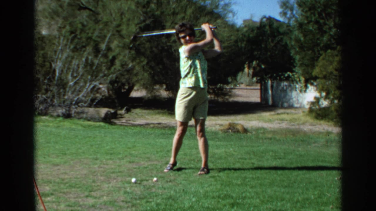 Woman in sleeveless patterned top swinging a golf club on a sunny course.