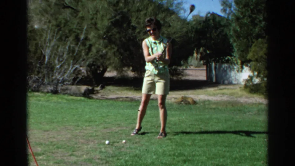 A woman in a patterned sleeveless top stands after swinging a golf club on a Scottsdale course in 1968 home-movie footage.