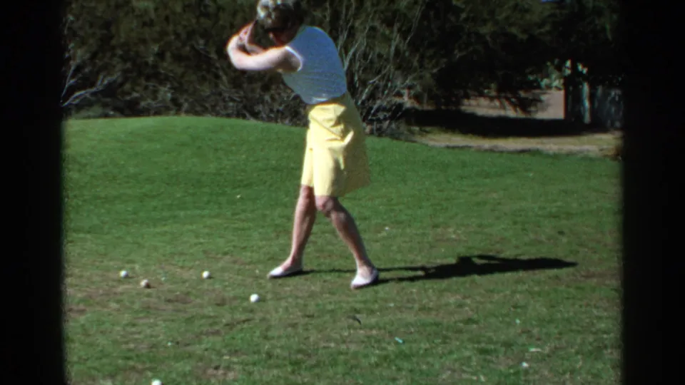 A woman golfer in 1968 Scottsdale follows through during practice, with golf balls scattered on the grass in front of her.