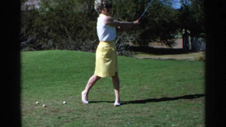 The woman golfer follows through on a sunlit Scottsdale course, with trees and scattered golf balls visible in the 1968 home movie frame.