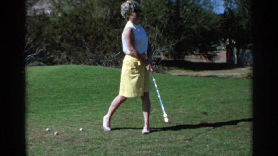 A woman in mid-century golf attire stands after a swing on a Scottsdale course, with golf balls on the grass and trees behind her.