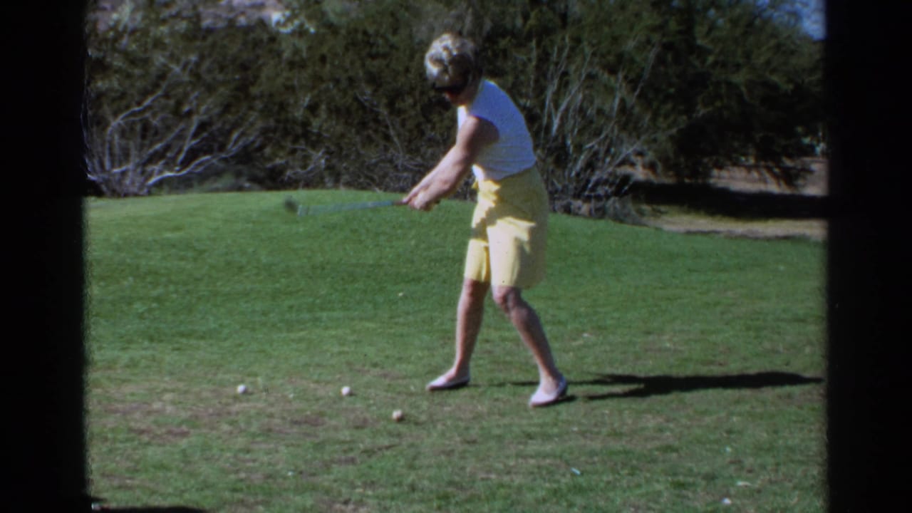 Woman golfer addresses a ball on a sunny grass course with desert shrubs behind her.