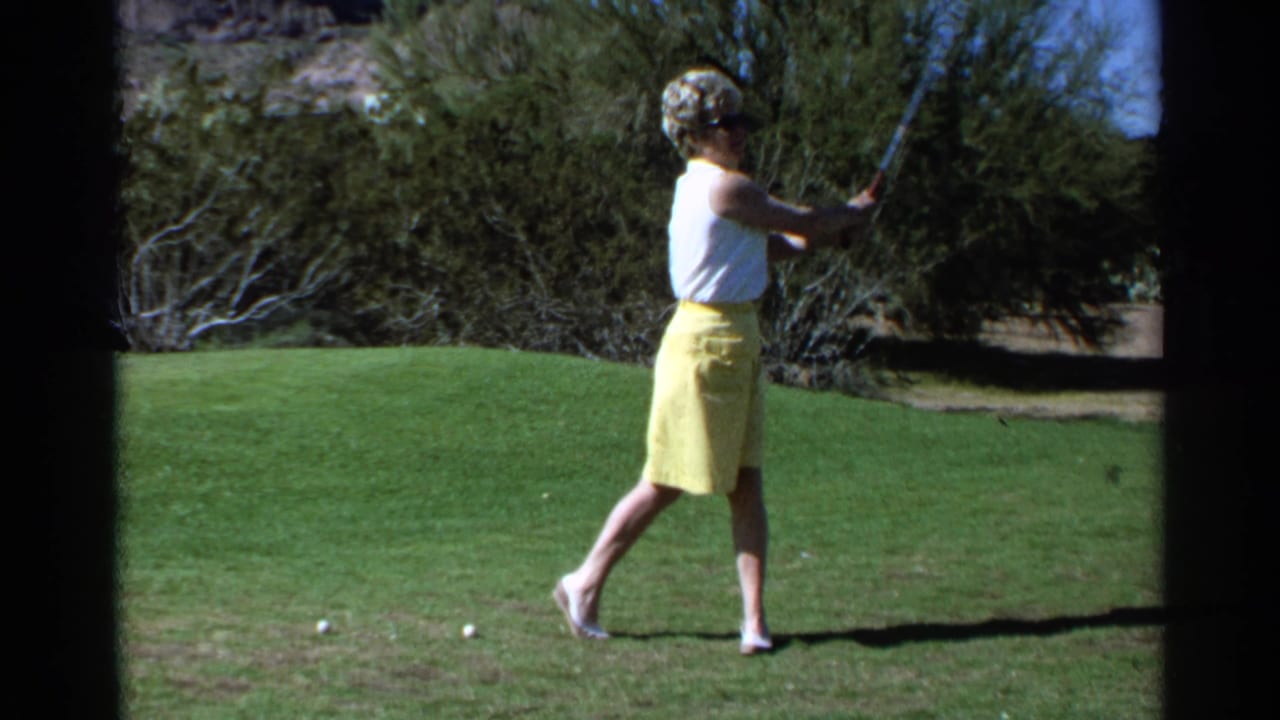 In 1968 Scottsdale, Arizona, a woman swings a golf club on a sunny green with desert shrubs behind her.