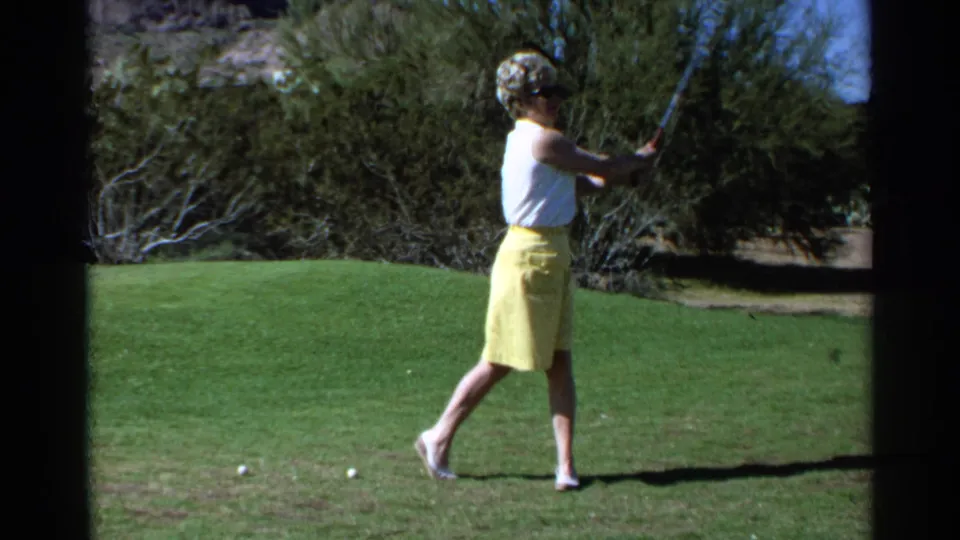 In 1968 Scottsdale, Arizona, a woman swings a golf club on a sunny green with desert shrubs behind her.